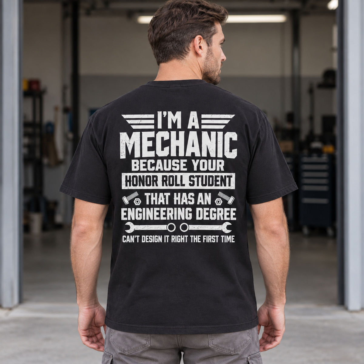 I'm A Mechanic engineering degree graphic tee in black, male model showing the back print outside a garage