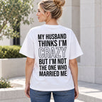 White funny wife graphic tee on a female model outdoors, showing the black My Husband Thinks I'm Crazy back print