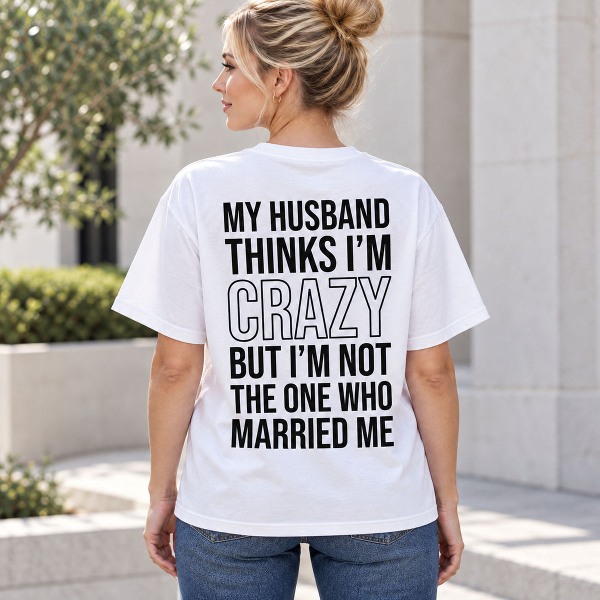 White funny wife graphic tee on a female model outdoors, showing the black My Husband Thinks I'm Crazy back print
