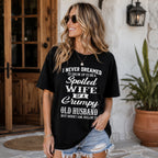 Spoiled Wife of a Grumpy Old Husband black front print graphic tee on a woman at a rustic doorway