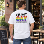I'm Not Gay But My Boyfriend Is white graphic tee, back print with black text and rainbow GAY lettering on a model outside a cafe