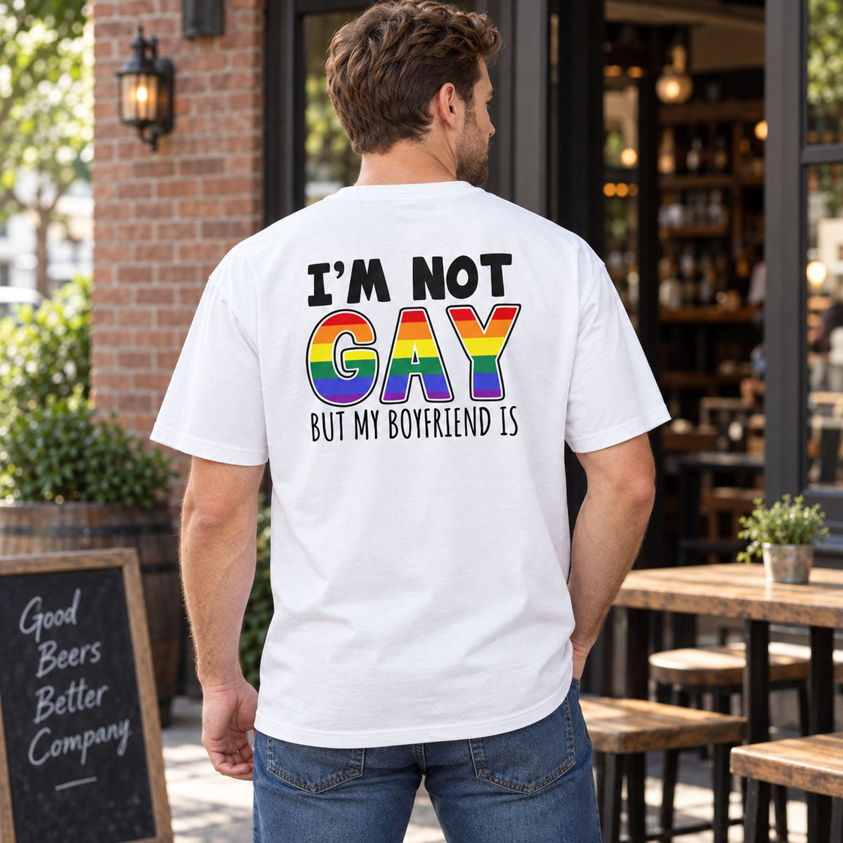 I'm Not Gay But My Boyfriend Is white graphic tee, back print with black text and rainbow GAY lettering on a model outside a cafe