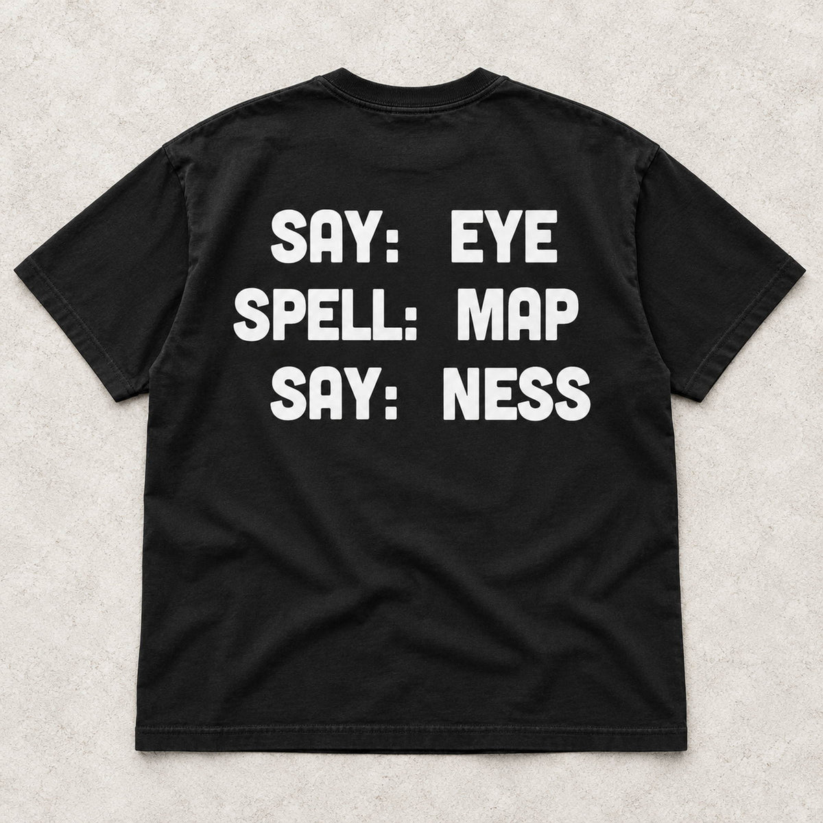 Say Eye Spell Map Say Ness white lettering on a black T-shirt back flat lay on a textured surface