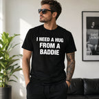 Male model wearing black I Need a Hug From a Baddie funny front-print graphic tee in a bright living room
