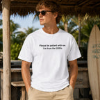 Male model wearing white Please Be Patient I'm From the 1900s funny front-print graphic tee at a tropical surf setting