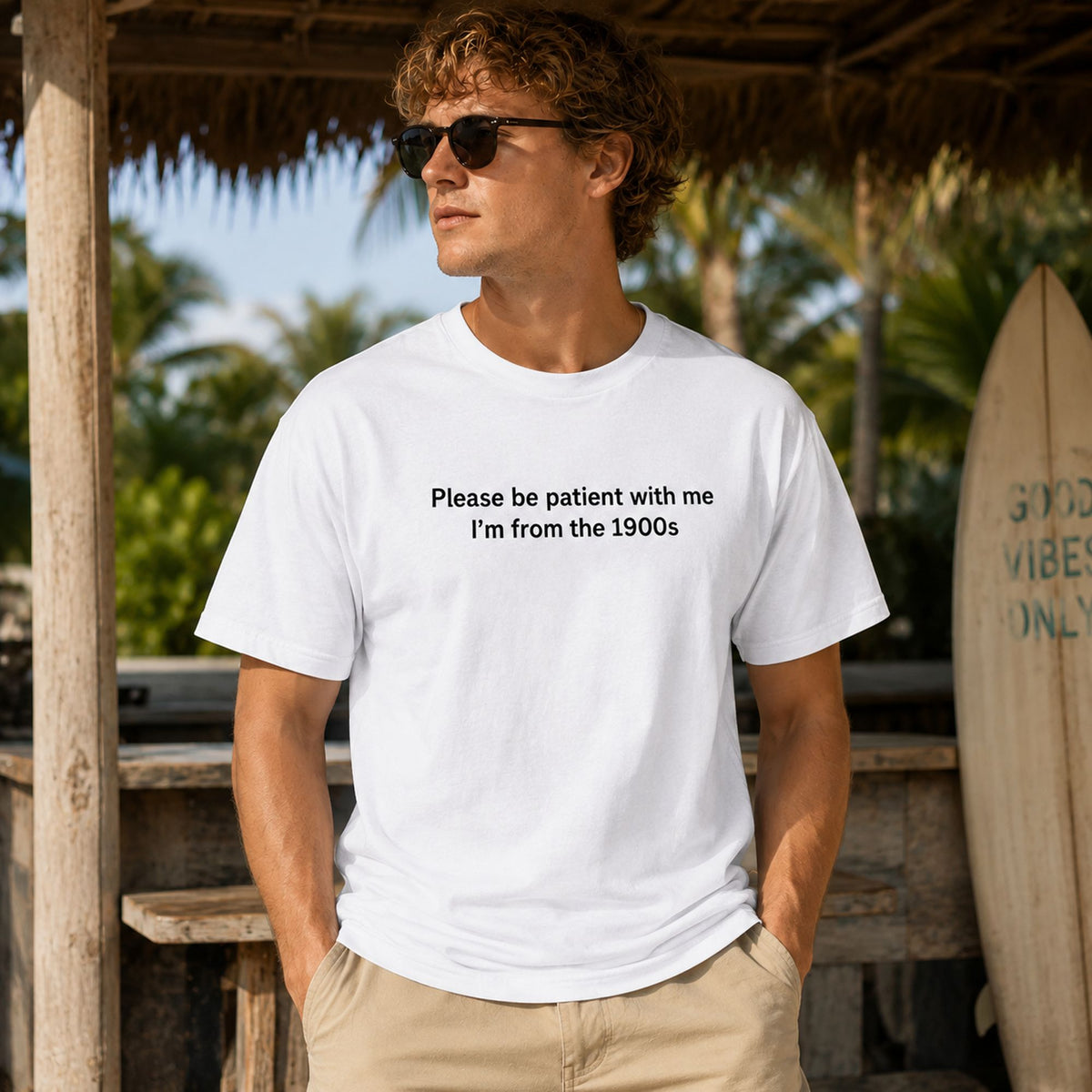 Male model wearing white Please Be Patient I'm From the 1900s funny front-print graphic tee at a tropical surf setting